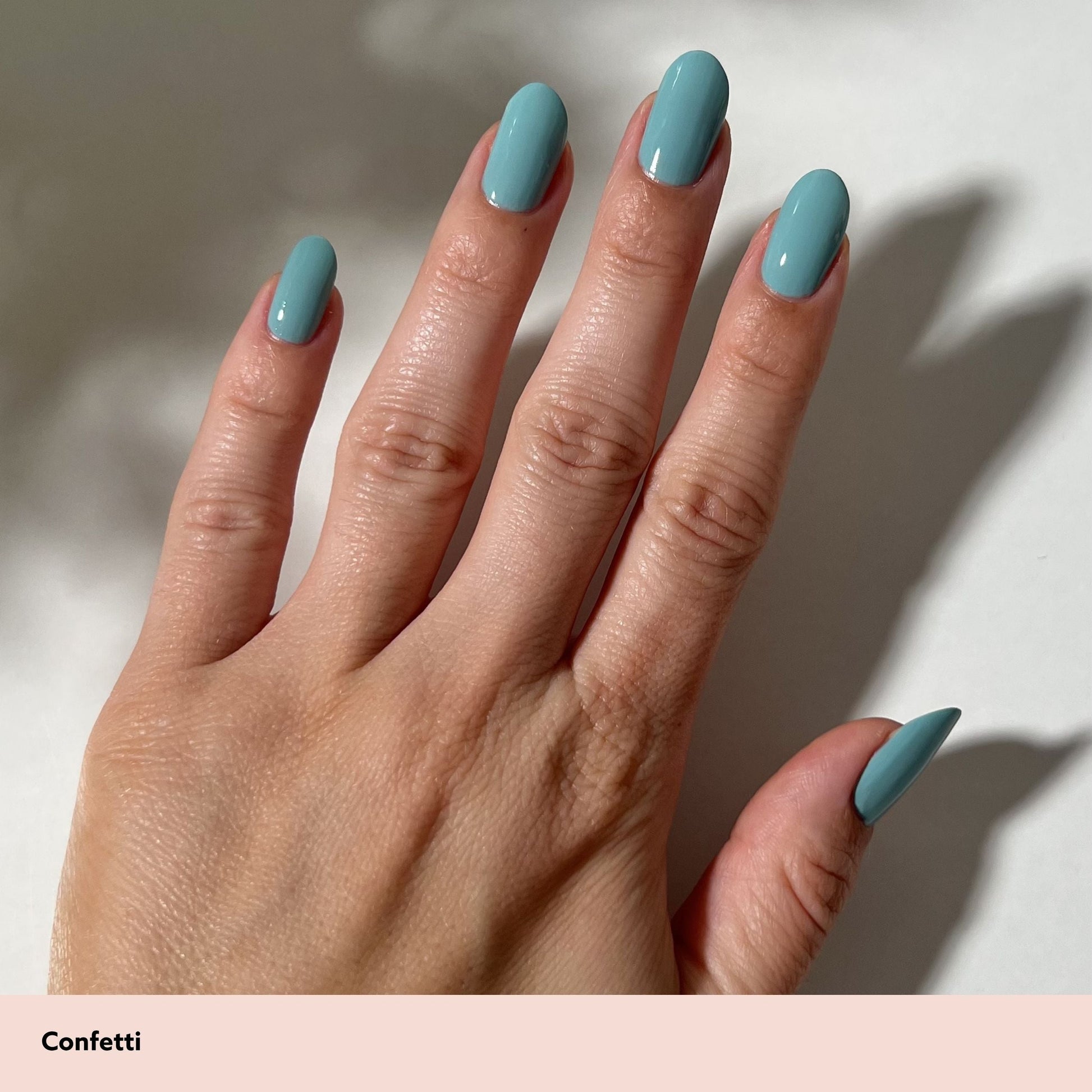 A close-up of a hand with almond-shaped nails painted in a dusty light blue shade, labeled 'Confetti' on a pale pink strip at the bottom. The background is a clean white surface with soft shadows.