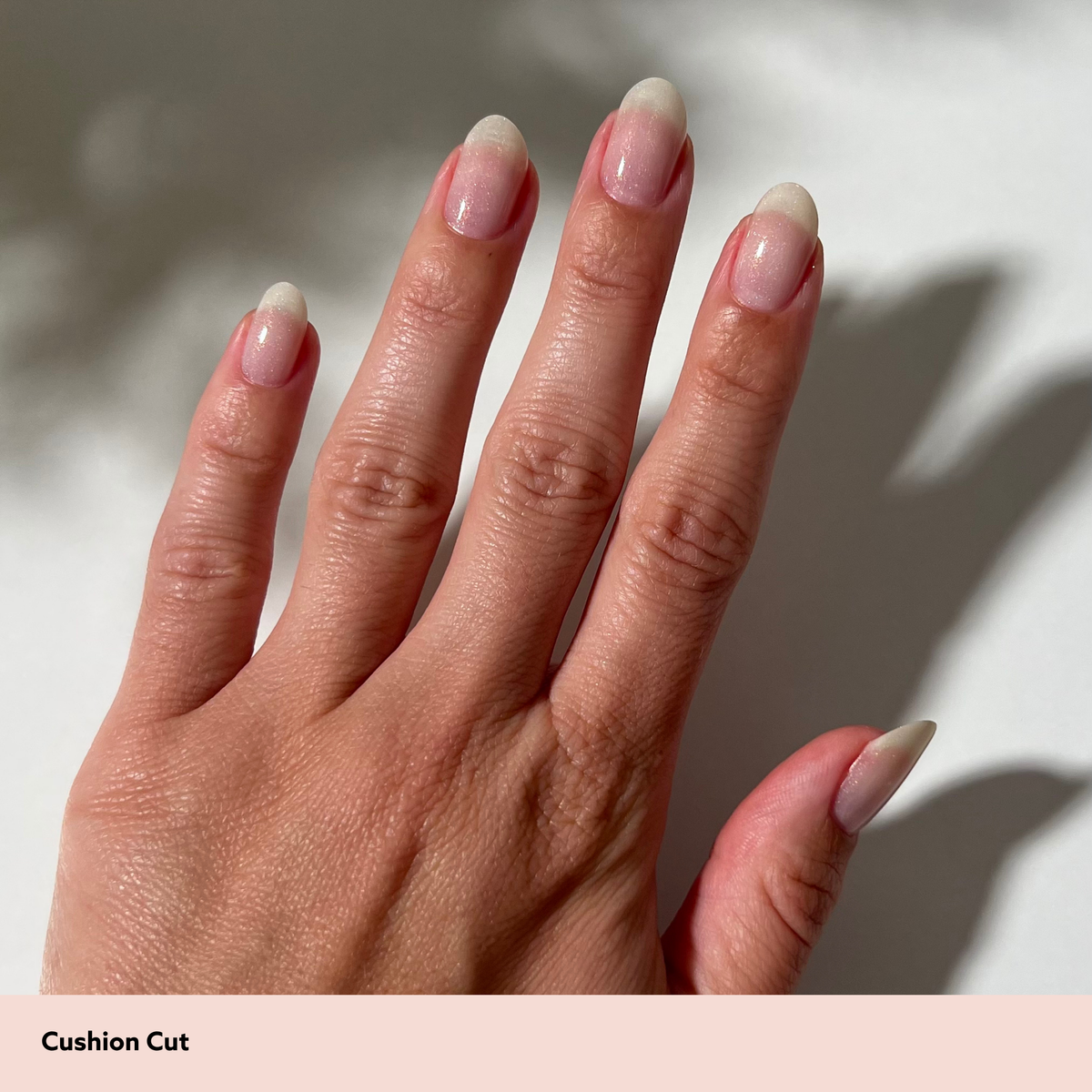 A close-up of a hand showing a cushion cut manicure with a soft pink-to-nude gradient on the nails, set against a light background with soft shadows. A beige banner at the bottom reads 'Cushion Cut'.