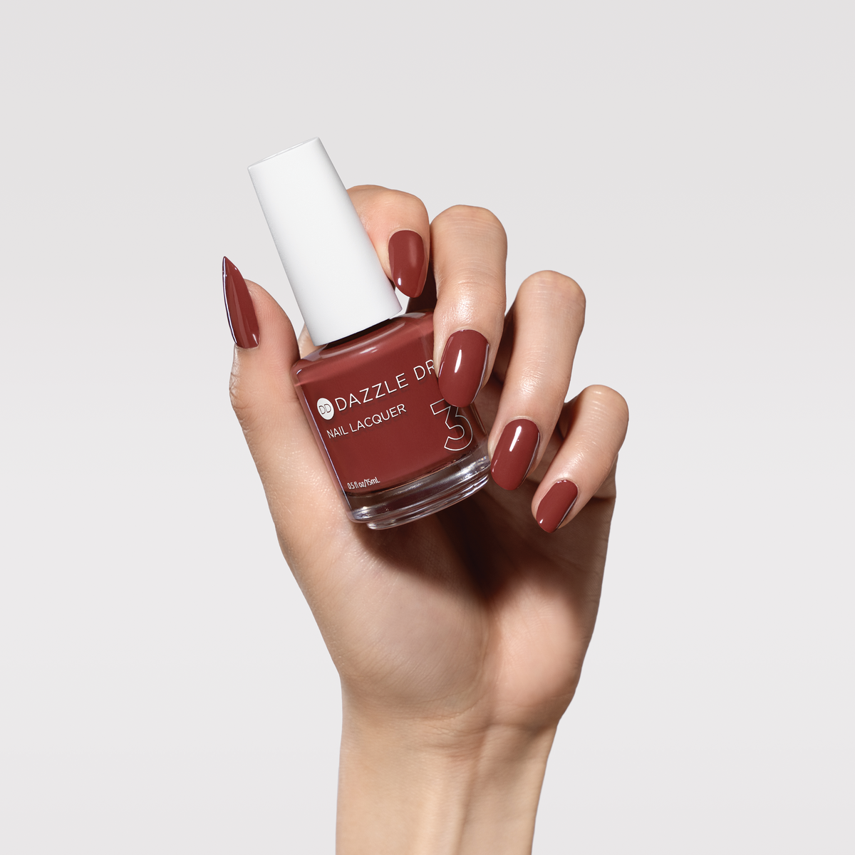 A hand holding a bottle of Dazzle On nail lacquer in a dusty rose-brown shade, with the nails painted to match, against a light gray background.
