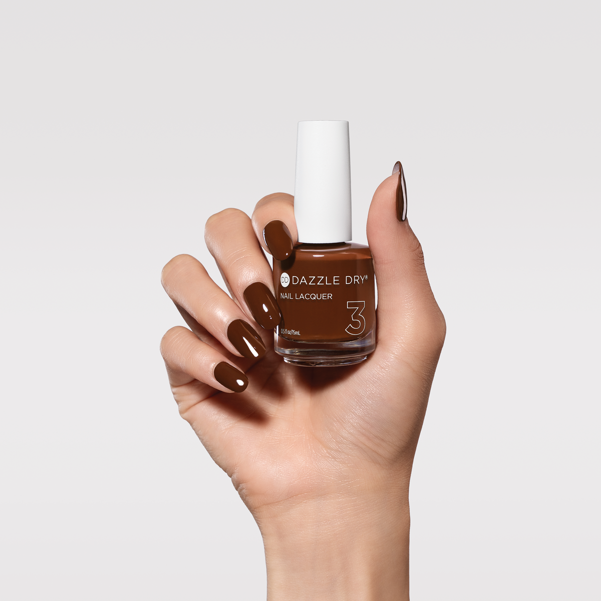A hand holding a bottle of Dazzle Drop 3 nail lacquer, showing a glossy dark brown nail polish color applied to the fingernails, against a light gray background.