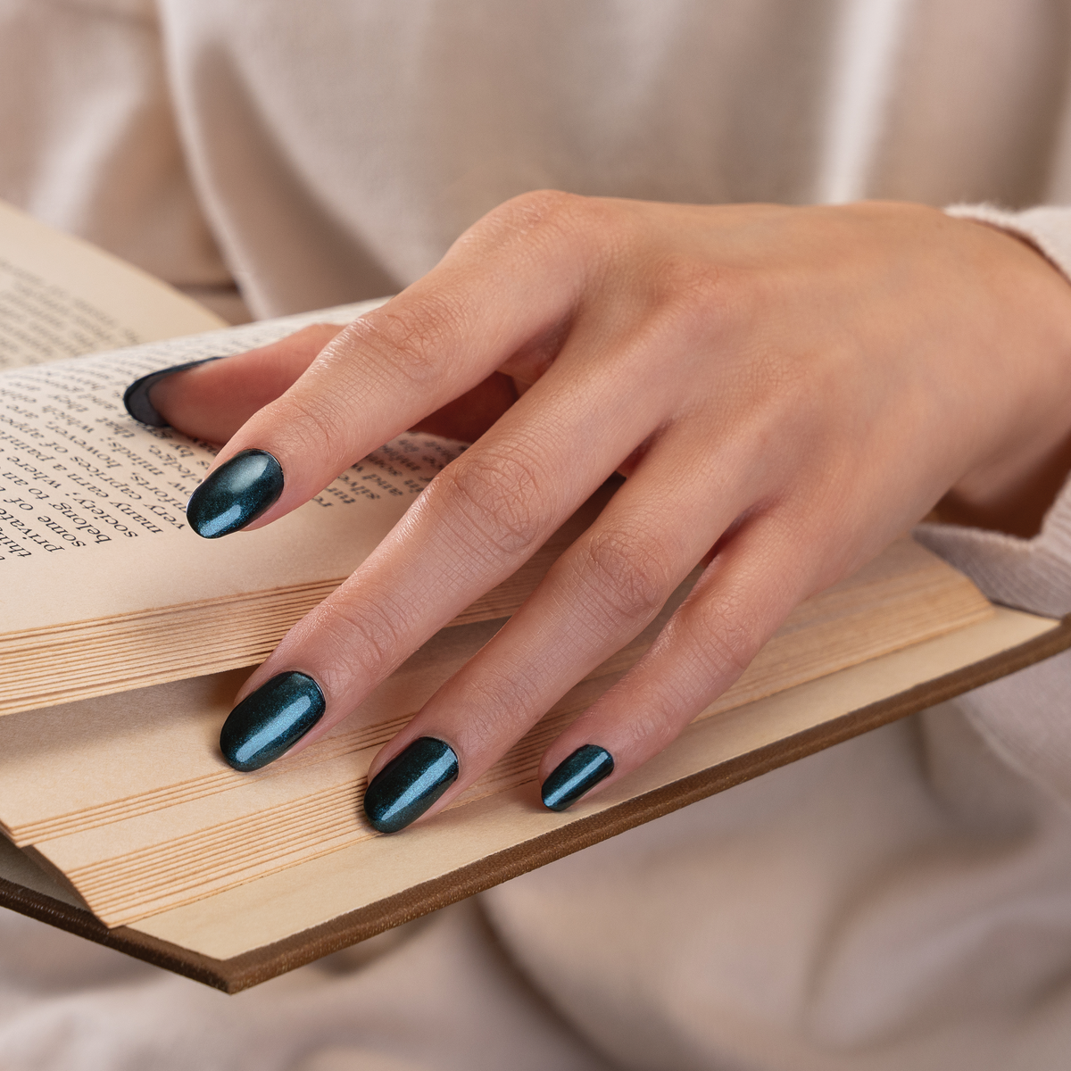 A close-up of a hand with glossy dark teal manicured nails resting on an open book, wearing a soft beige knit sweater.