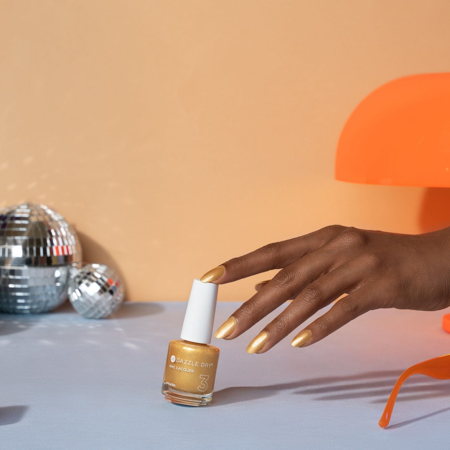 Hand with gold nail polish holding a bottle of gold nail polish against an orange background