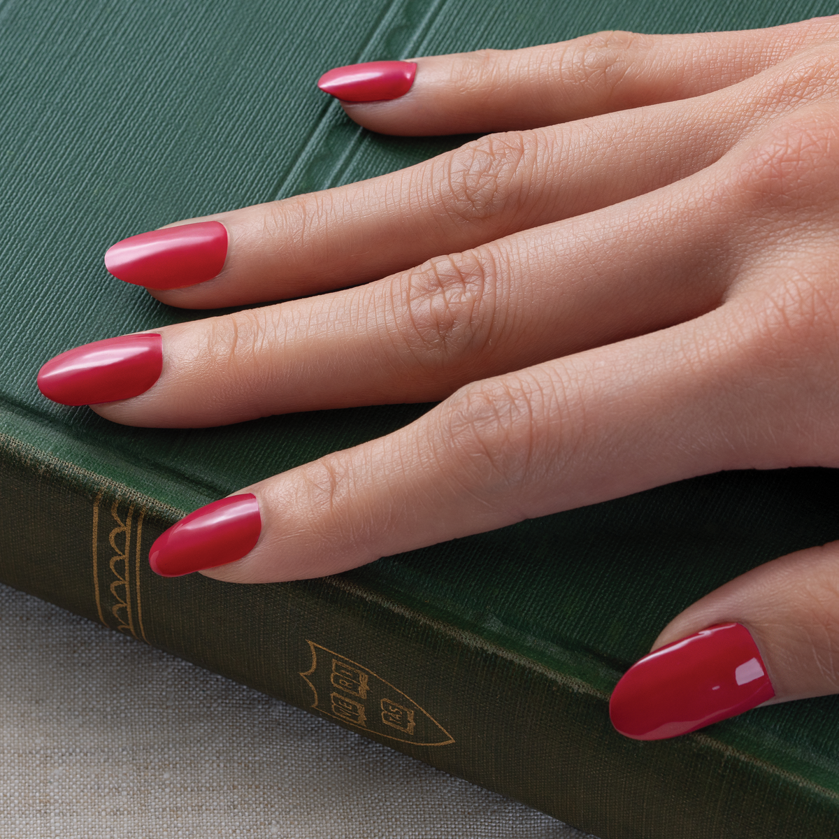 A close-up of a hand with long, glossy red manicured nails resting on a textured green book.
