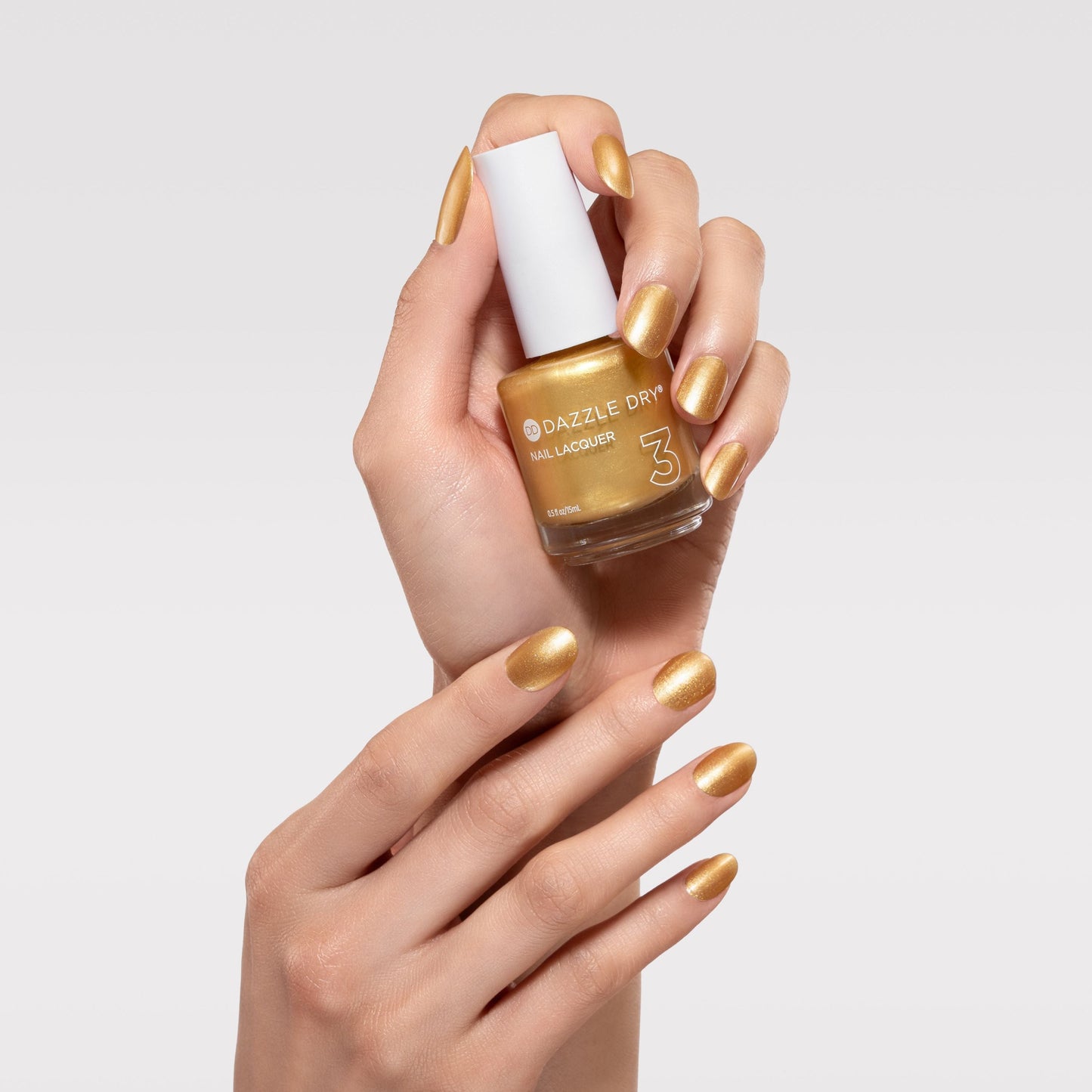 Hand with gold nail polish holding a bottle of nail polish against a light gray background