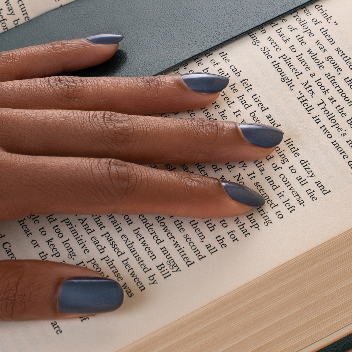 A close-up of a hand with medium skin tone and muted slate blue nail polish resting on an open book with printed text.