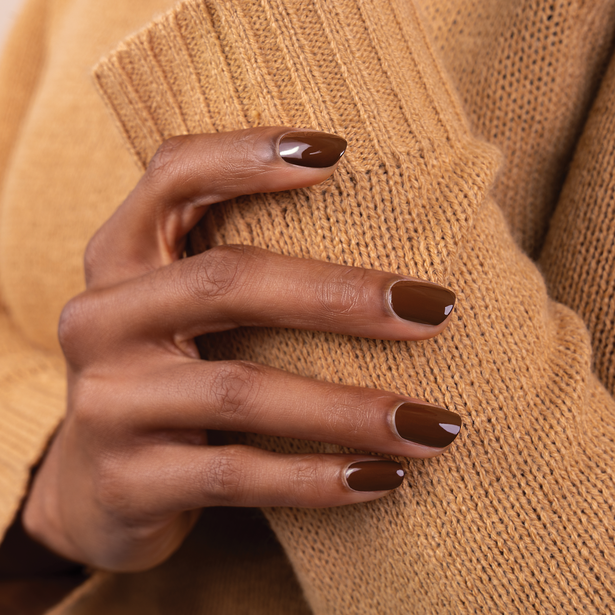 A close-up of a hand with glossy dark brown manicured nails resting on a ribbed camel-colored knit sweater.
