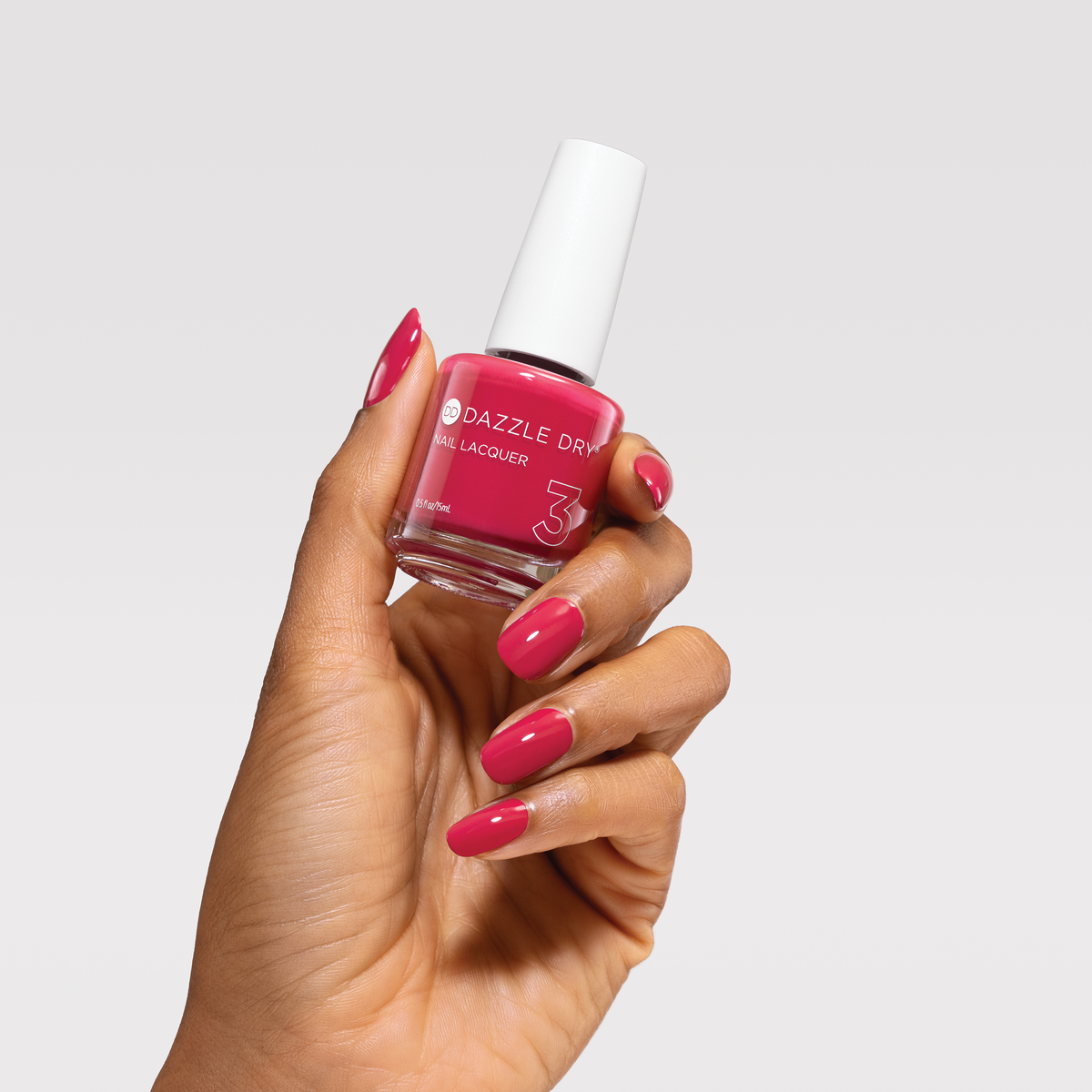 A hand with bright pink nail polish holding a bottle of Dazzle Dye nail lacquer in the same vibrant pink shade, set against a solid light gray background.