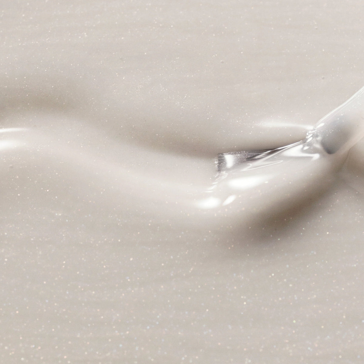A close-up of a glossy, shimmering cream-colored cosmetic cream with fine silver glitter, swirled on a matching background with a small brush visible in the upper right.