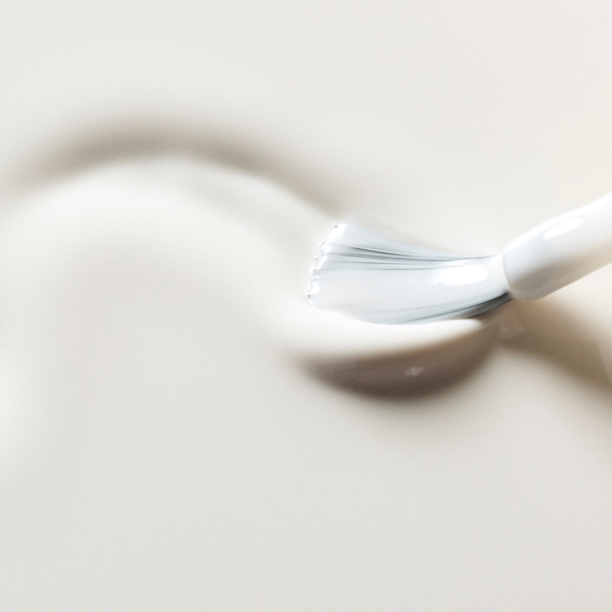 A close-up of a white cosmetic brush applying or spreading a thick, creamy white product on a smooth, light beige surface.