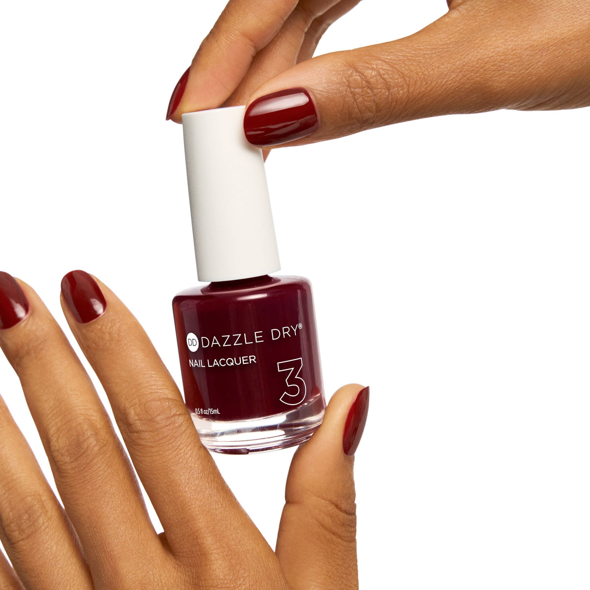 A bottle of Dazzle Dry Nail Lacquer in a deep burgundy shade, held by a hand with matching burgundy manicured nails against a solid white background.