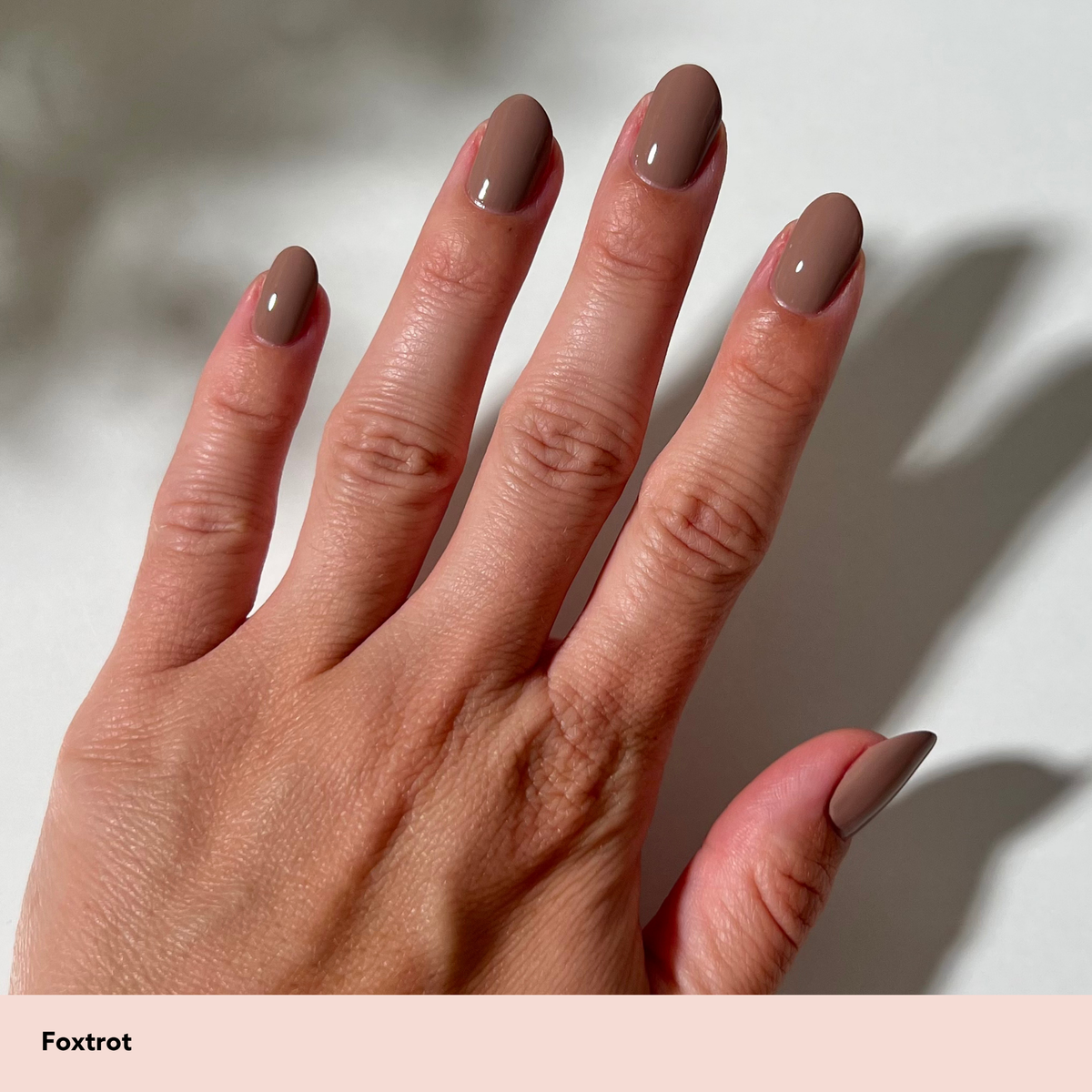 A close-up of a hand with long, almond-shaped nails painted in a glossy taupe-brown shade labeled 'Foxtrot' on a pale pink strip at the bottom. The hand is shown against a light background with soft shadows.