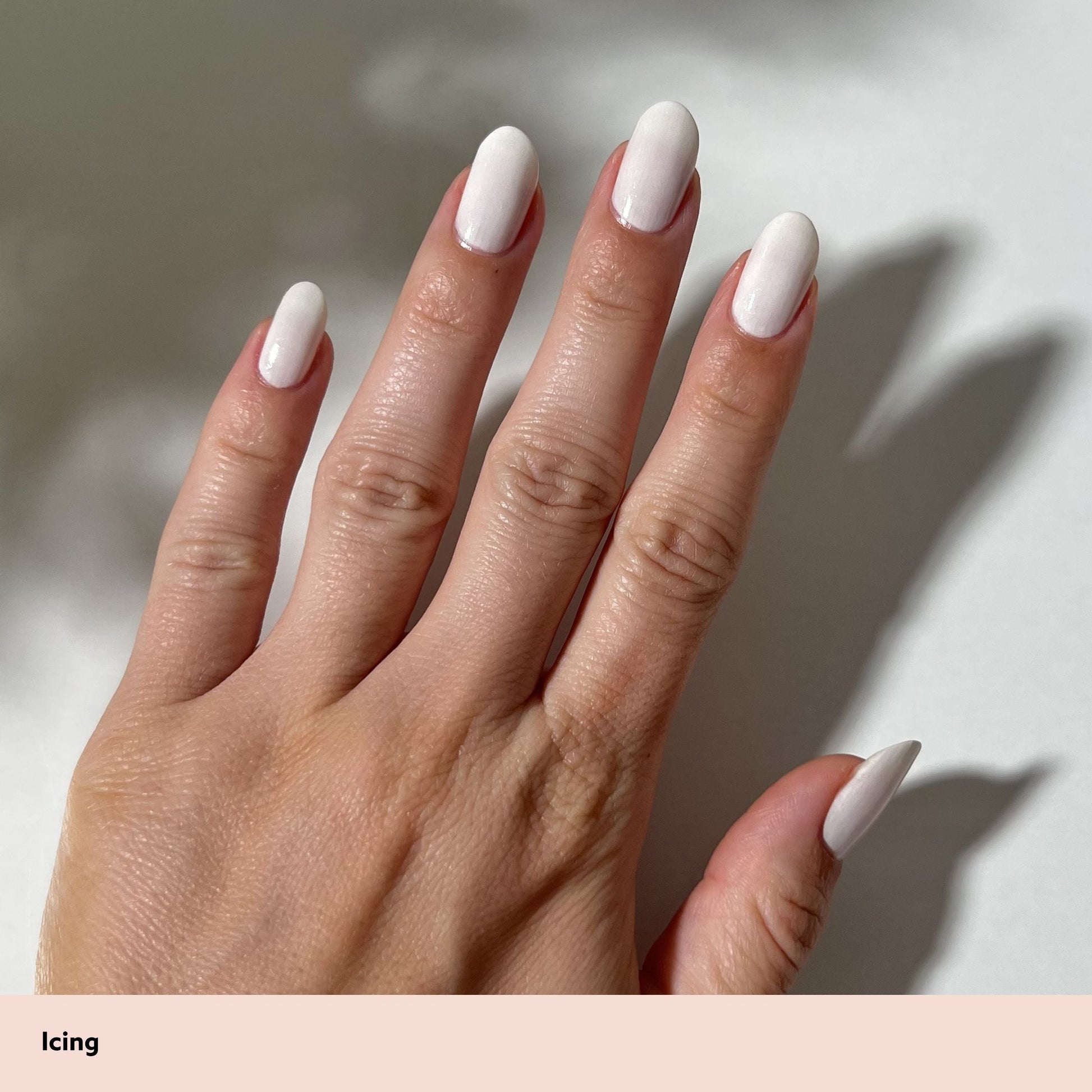 A close-up of a hand with long, almond-shaped nails painted in a glossy white 'Icing' manicure, set against a neutral light gray background with soft shadows. A pale pink label at the bottom left reads 'Icing'.