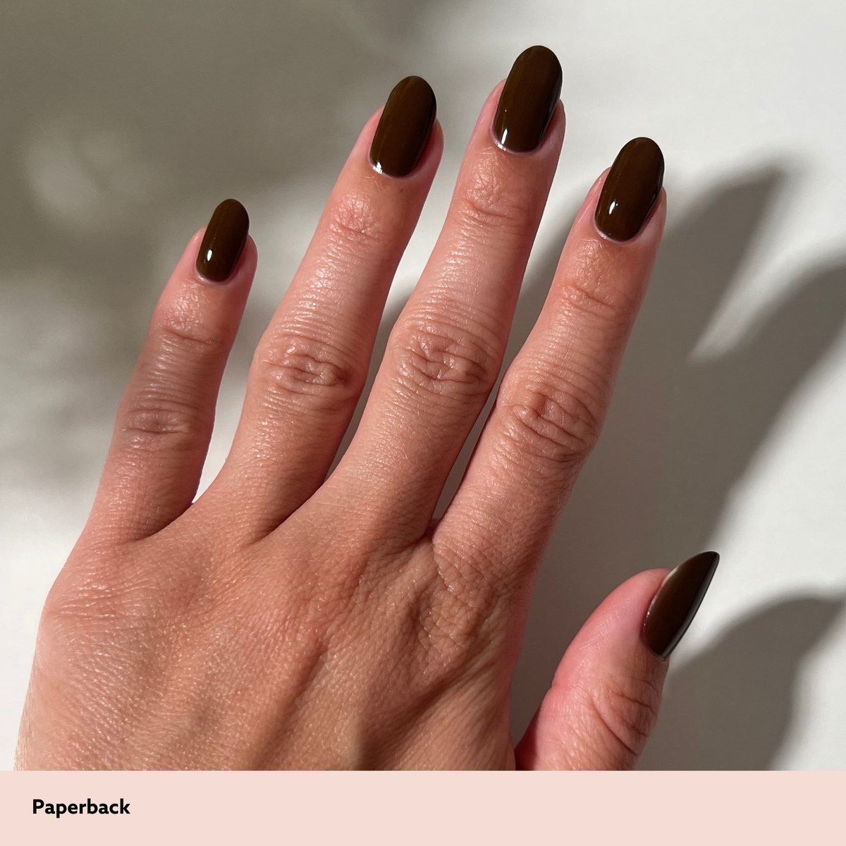 A close-up of a hand with glossy dark brown manicured nails, shown against a light neutral background with a soft shadow. A pale pink label at the bottom reads 'Paperback'.