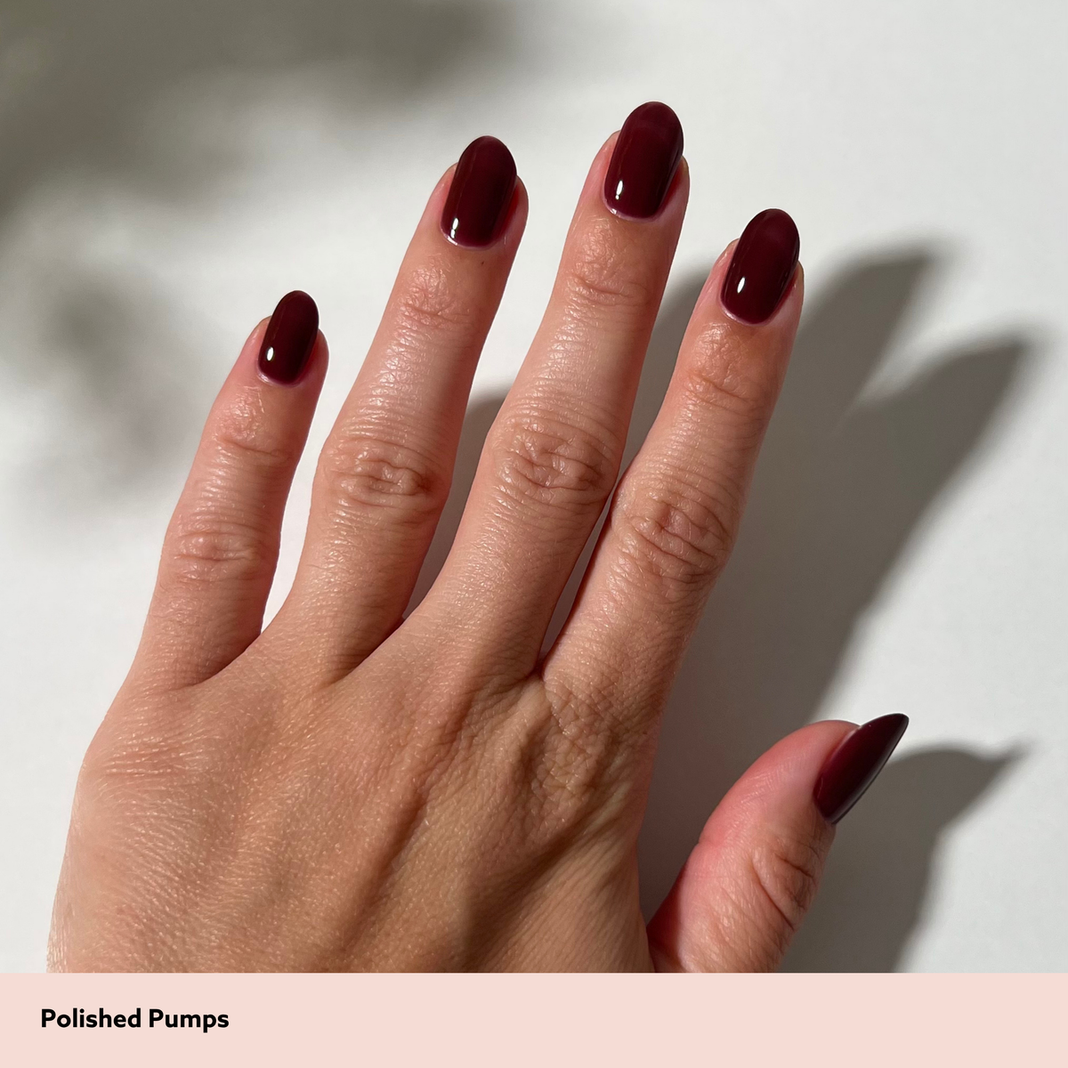 A close-up of a hand with long, almond-shaped nails painted in a deep glossy burgundy shade, with a text label at the bottom reading 'Polished Pumps'.