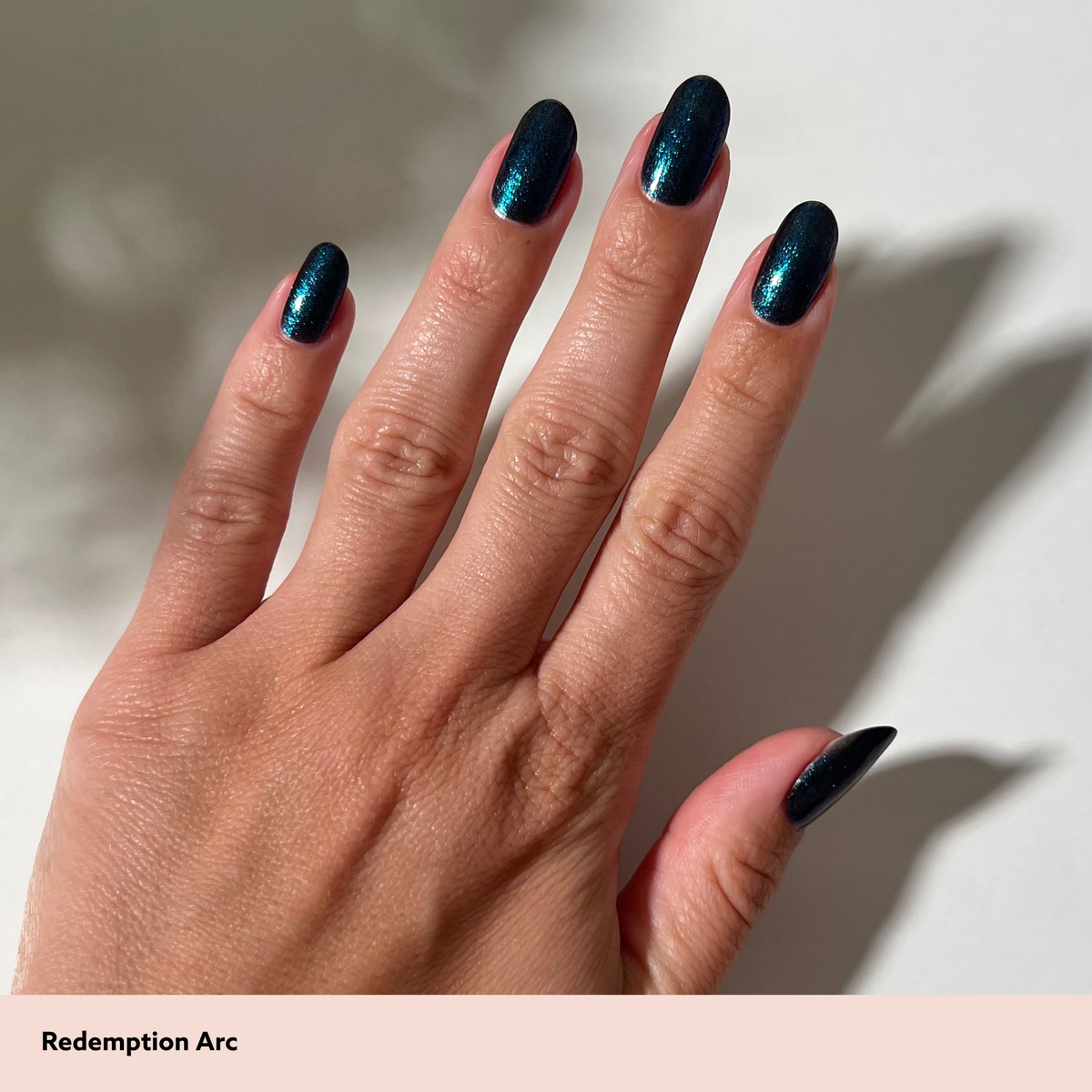 A close-up of a hand with almond-shaped nails painted in a deep metallic teal-blue nail polish, shown against a light neutral background with soft shadows. Text at the bottom reads 'Redemption Arc'.