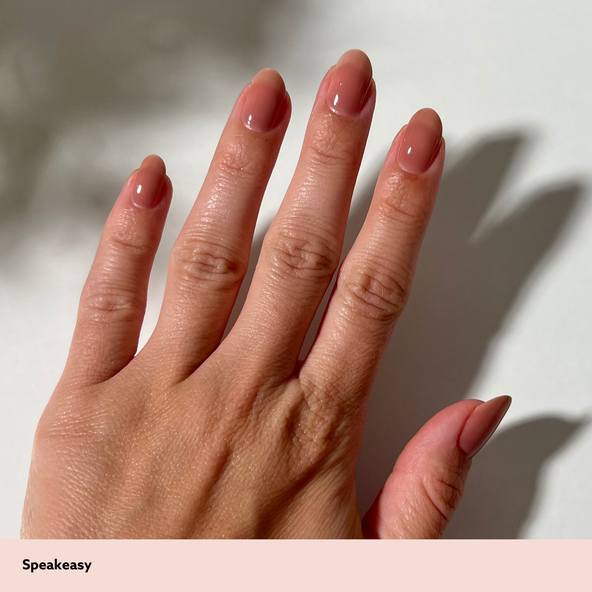 A close-up of a hand with neatly manicured nails painted in a glossy dusty pink shade, set against a light background with a soft shadow. The brand name 'Speakeasy' is printed in black text on a pale pink banner at the bottom left.