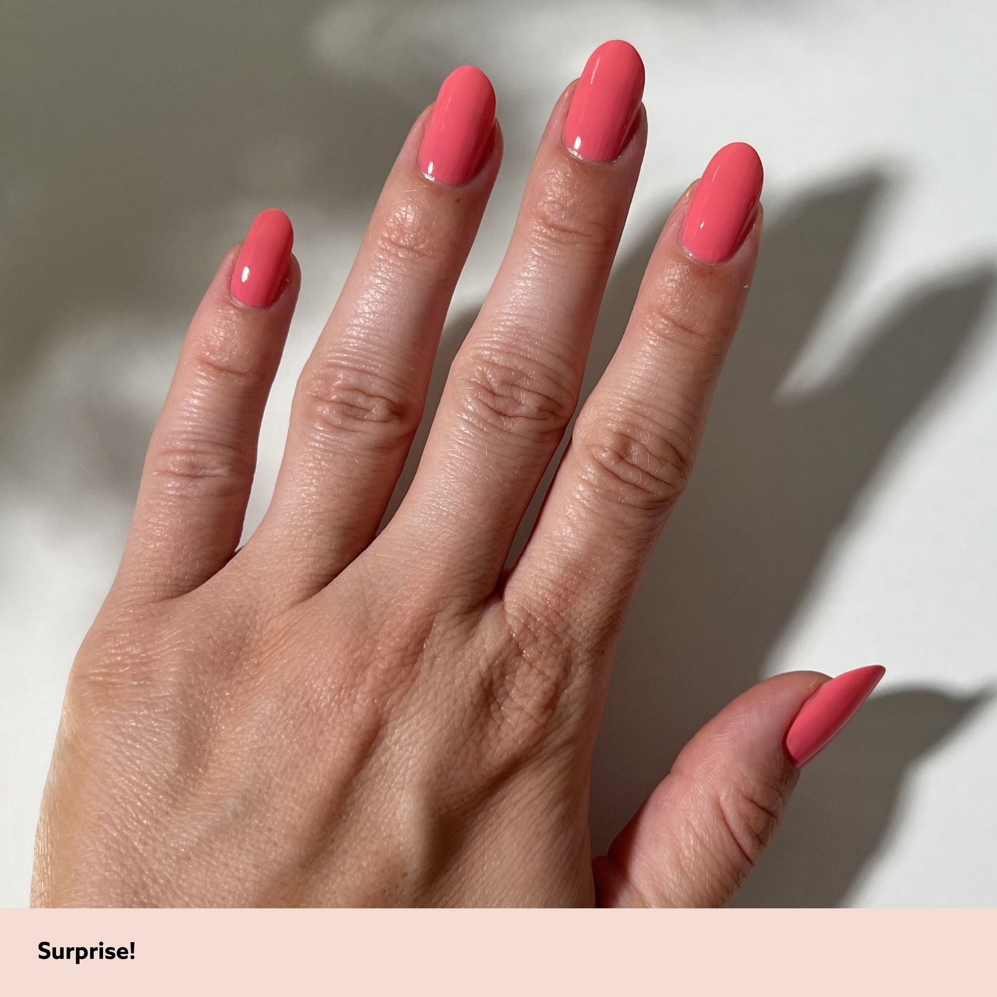 A close-up of a hand with long, almond-shaped nails painted in a solid coral pink color, set against a light background with soft shadows. A pale pink banner at the bottom contains the text 'Surprise!'.